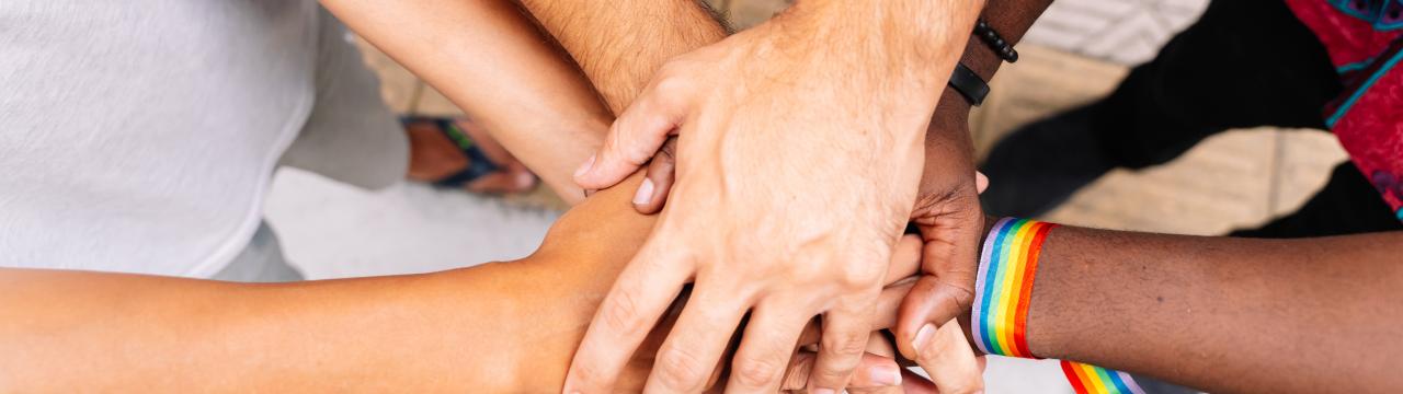 Mehrere unterschiedliche Hände übereinander gefaltet, eine Hand trägt dabei ein Symbol der LGBTQ-Bewegung. 